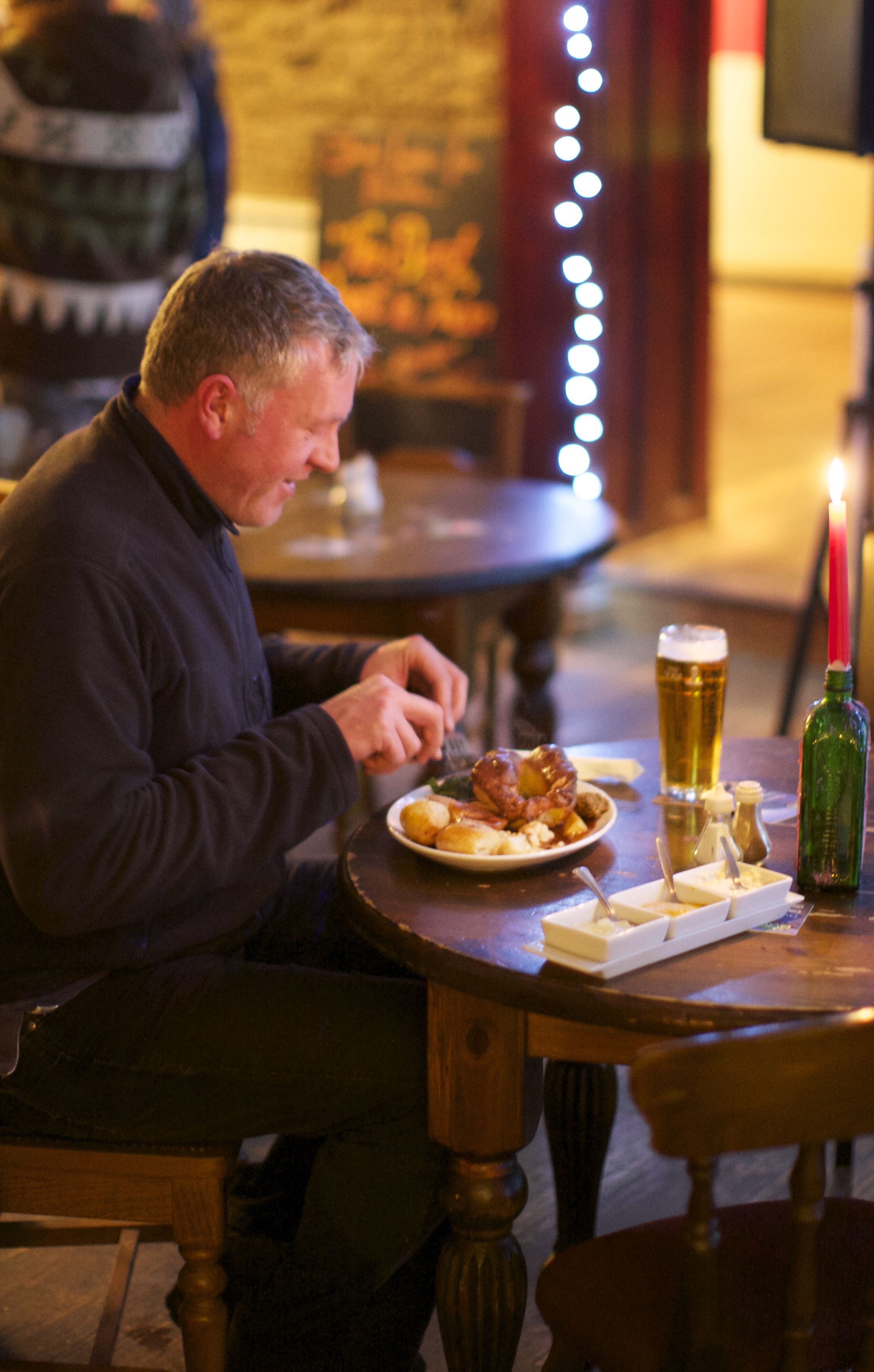 Un hombre sentado a una mesa de madera con un plato de comida, una jarra de cerveza y una vela.