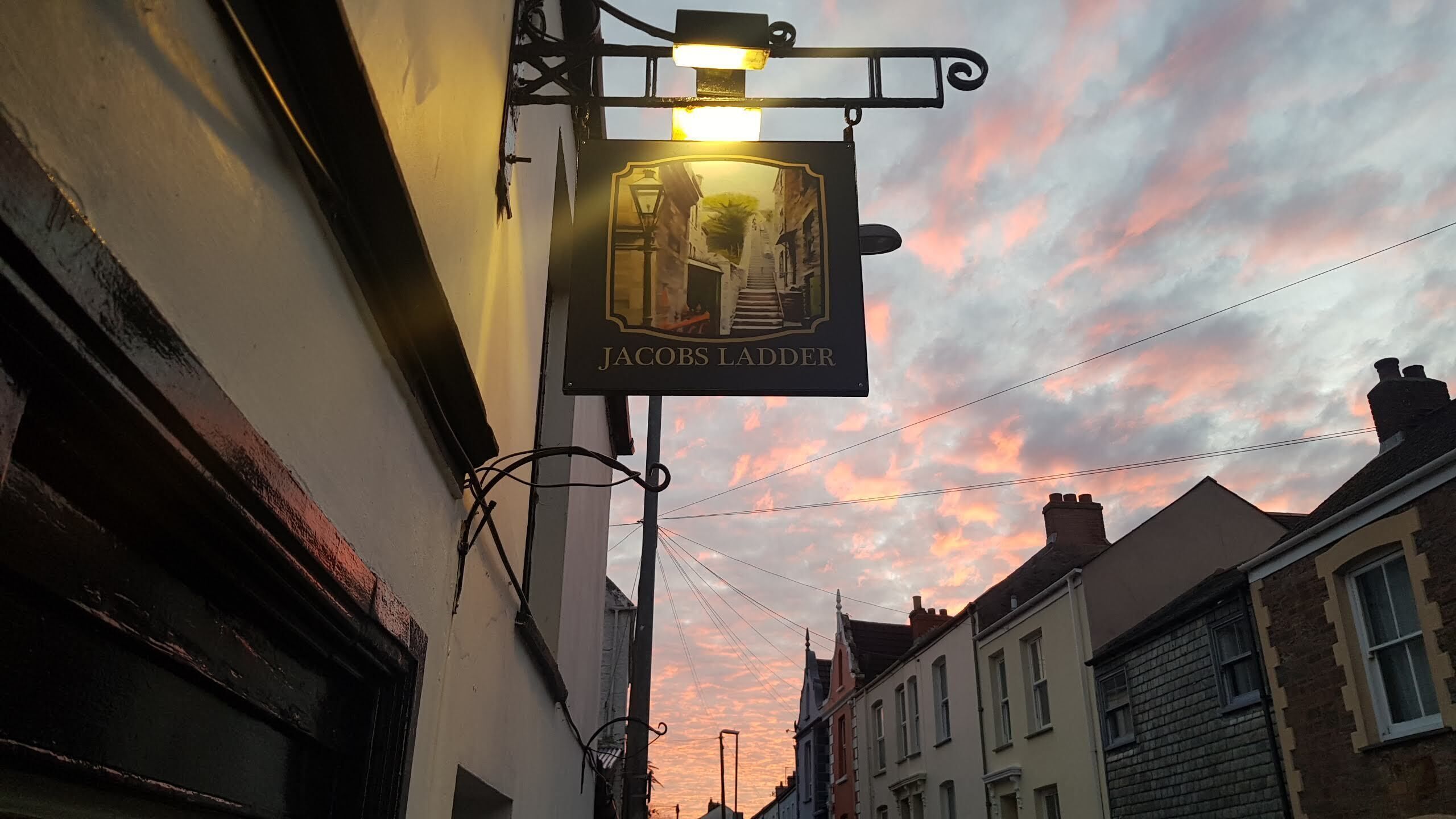 A street view with a pub sign reading "Jacob's Ladder" against a sunset sky.
