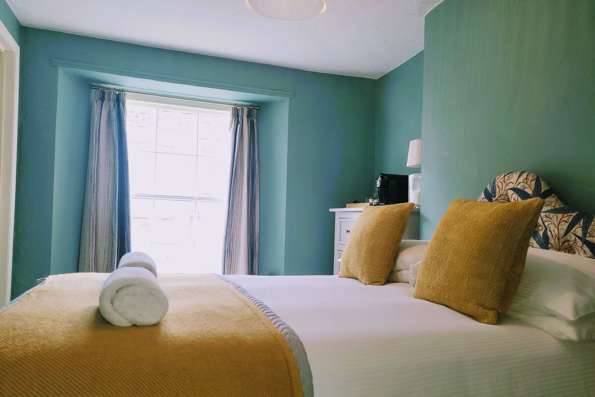 A hotel room with a bed, a window with curtains, a bedside table with a lamp, and a patterned pillow.