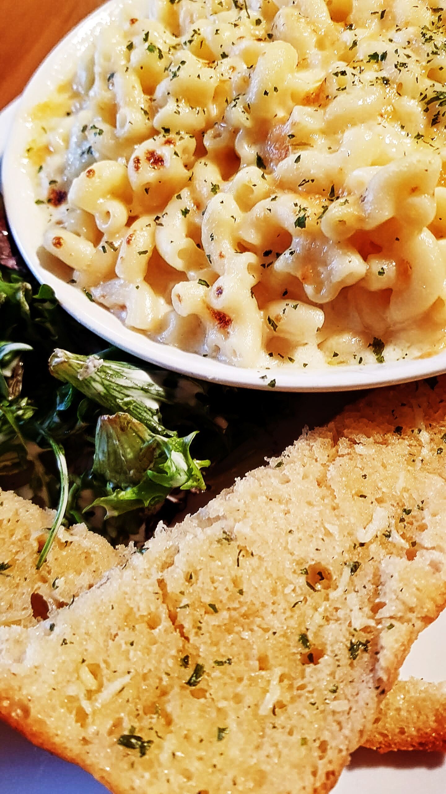 Plato de macarrones con queso, adornado con hierbas, acompañado de verduras de hoja y una rebanada de pan.
