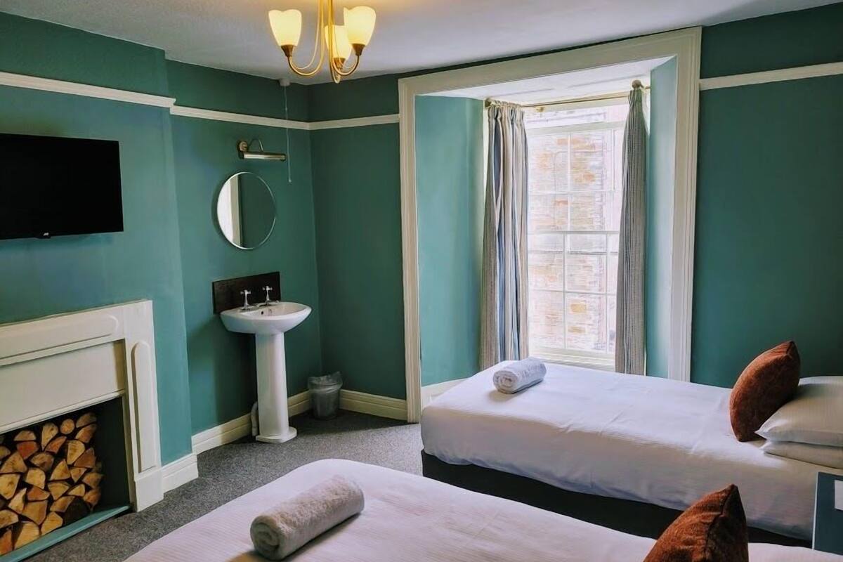 A hotel room with teal walls, a white sink, a mirror, a chandelier, a fireplace, two beds with pillows, and a window with curtains.