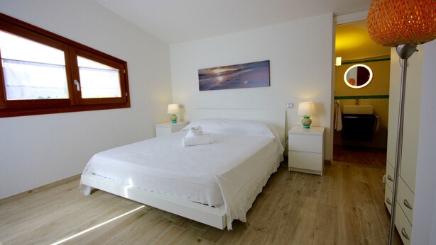 A bedroom with a large bed, two bedside tables with lamps, a painting on the wall, and a view of the bathroom through an open door. - Villa Franca (San Teodoro)