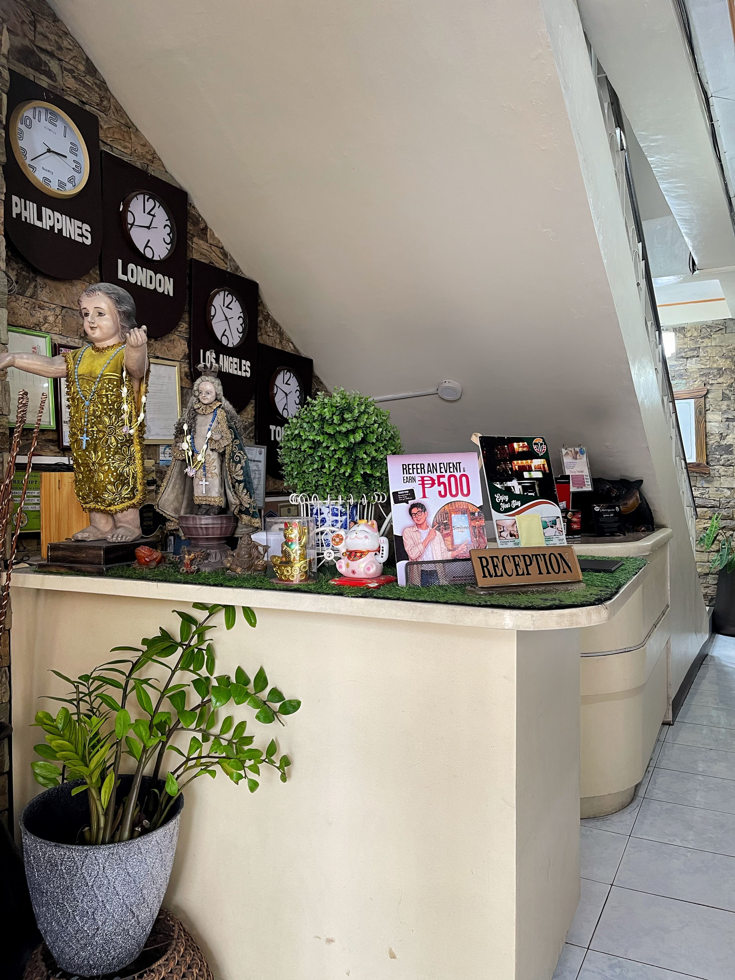 Un mostrador de recepción con un cartel que ofrece un bono de referencia, rodeado de relojes que marcan diferentes ciudades, una planta en maceta y varios objetos decorativos.