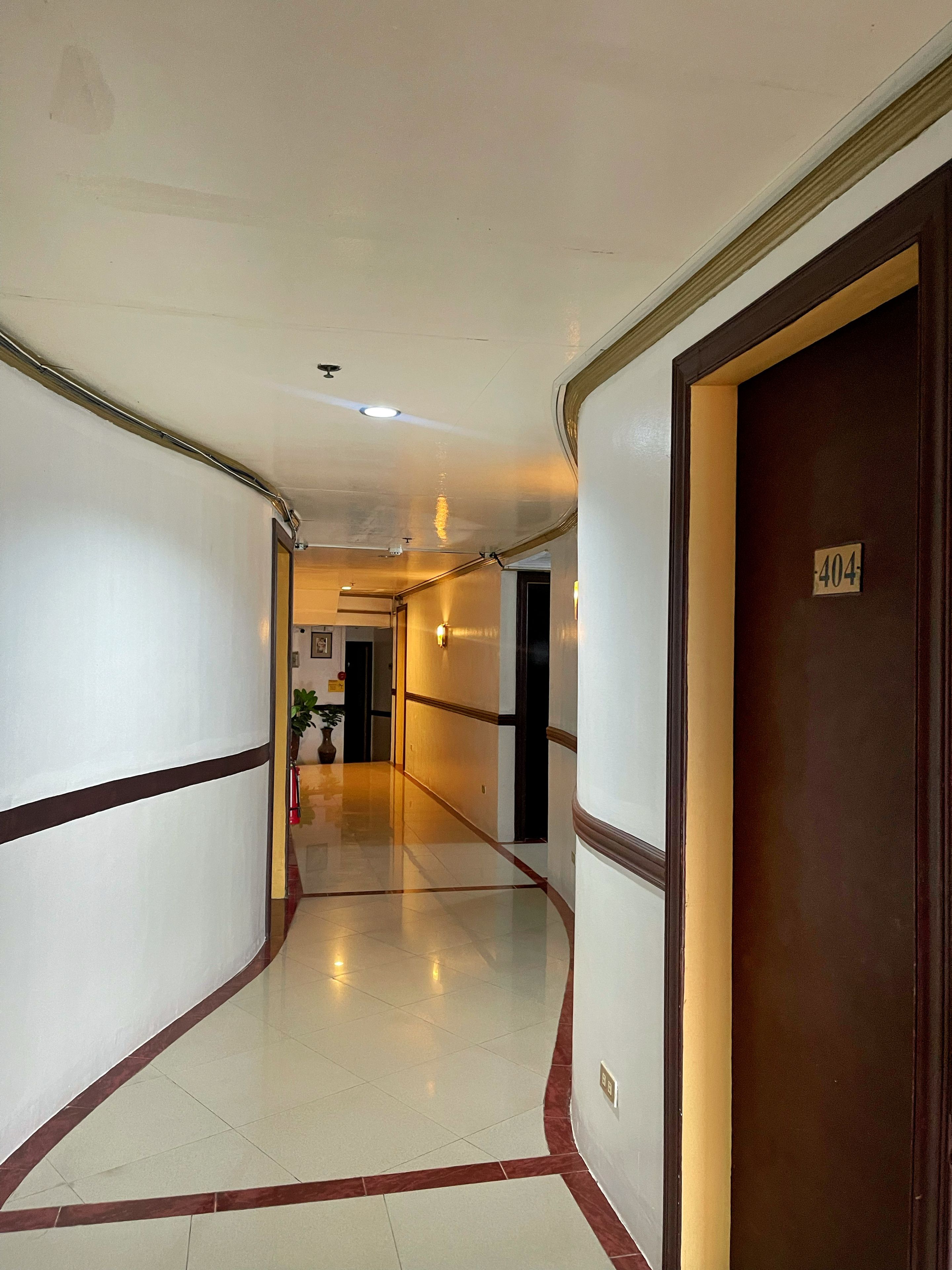 A hotel corridor with a wooden door marked '404' on the right, a white wall with a brown trim on the left, and a tiled floor.
