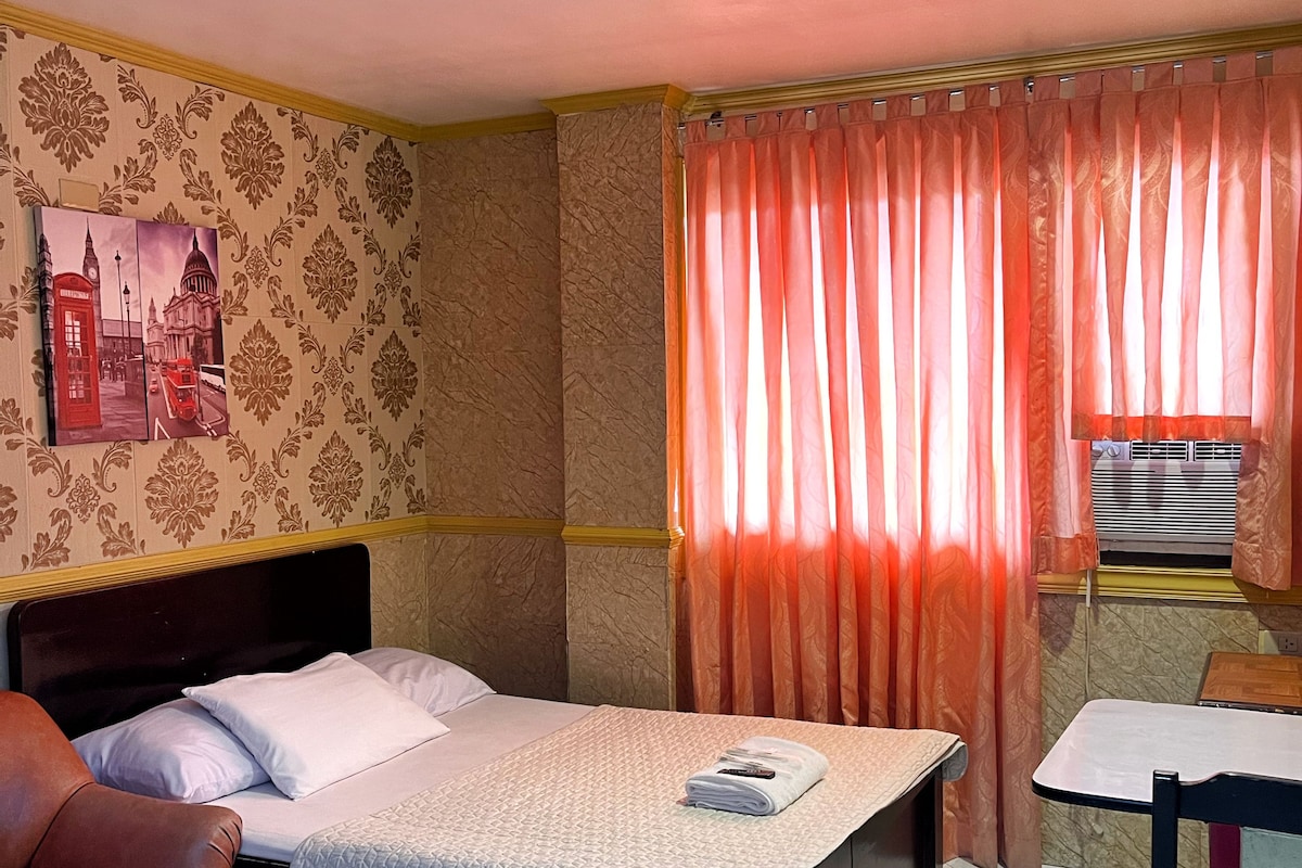 A hotel room with a bed, a chair, a table, a painting, and a window with curtains.