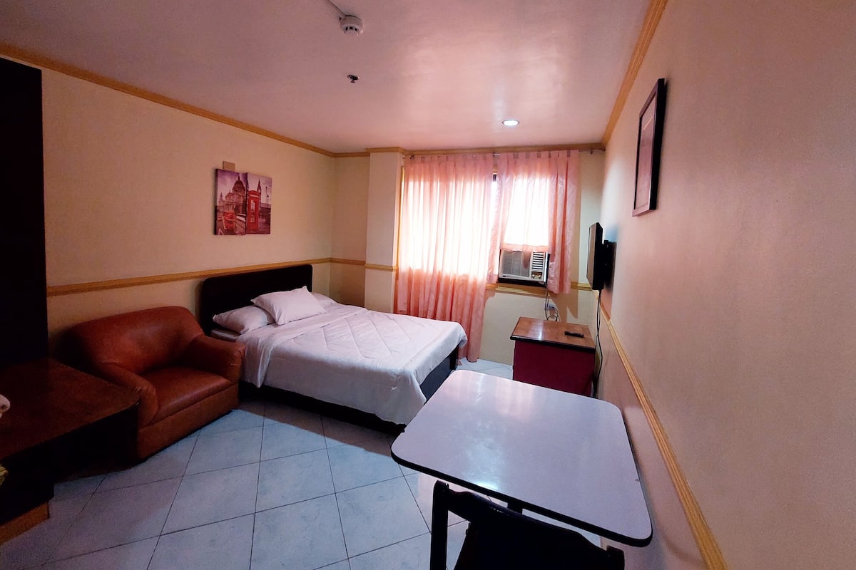 A hotel room with a bed, a red sofa, a small table, an air conditioning unit, and a television on the wall.