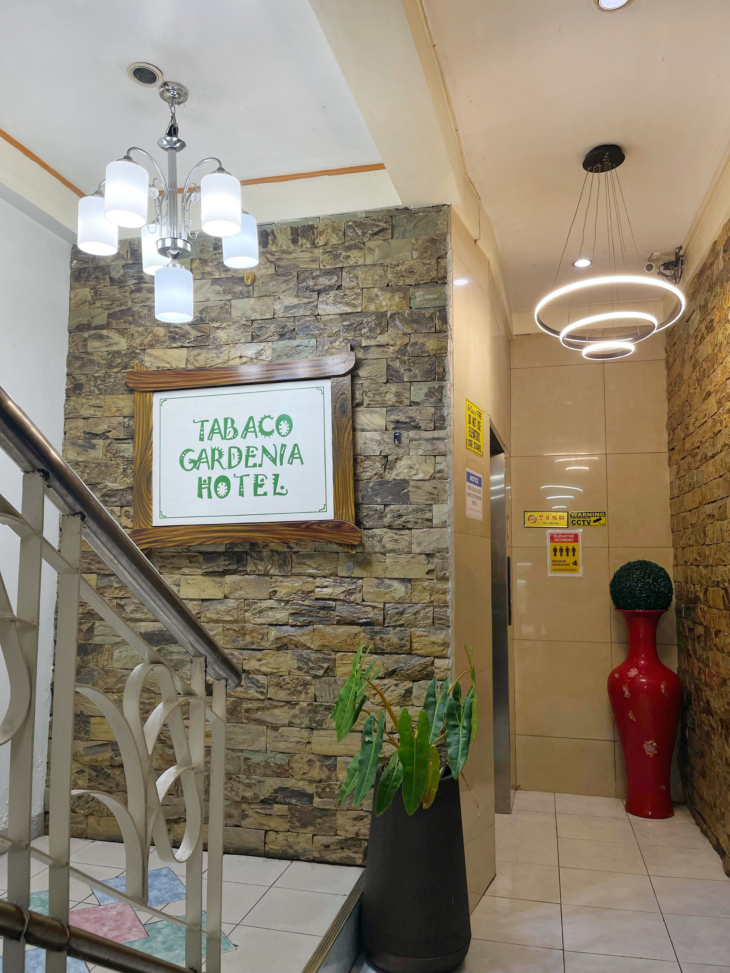 A hotel lobby with a staircase, a sign for 'Tabaco Gardenia Hotel', a potted plant, and a red vase.