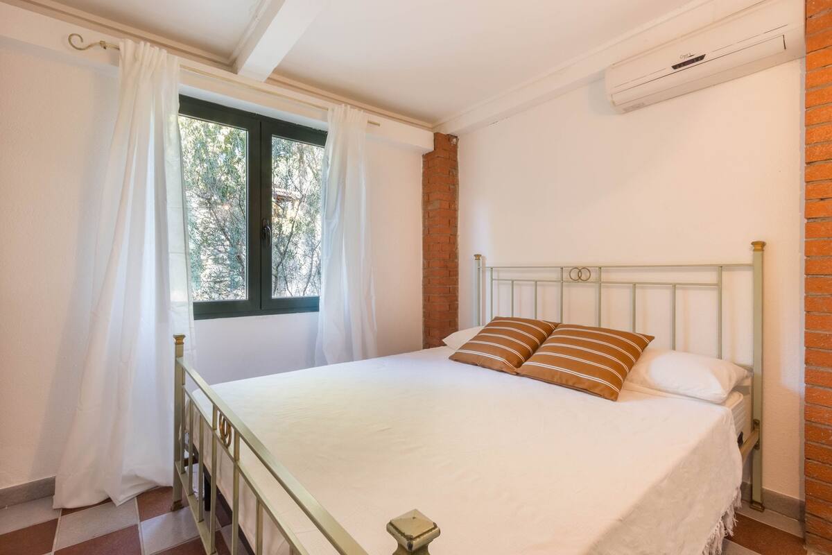 A bedroom with a bed, white curtains, a window, a brick wall, and an air conditioning unit.