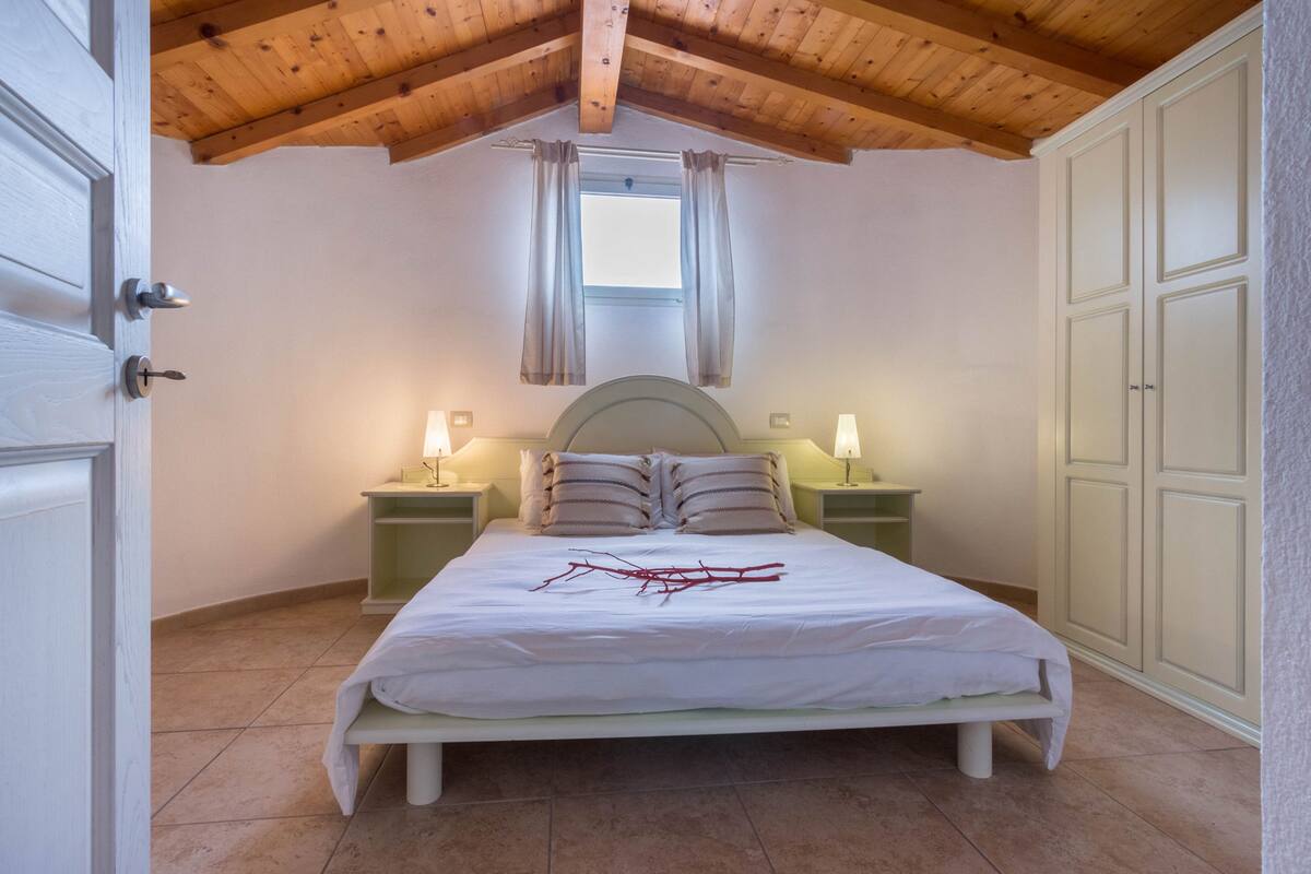 A bedroom with a wooden ceiling, a bed with white bedding, two bedside tables with lamps, and a window with curtains.