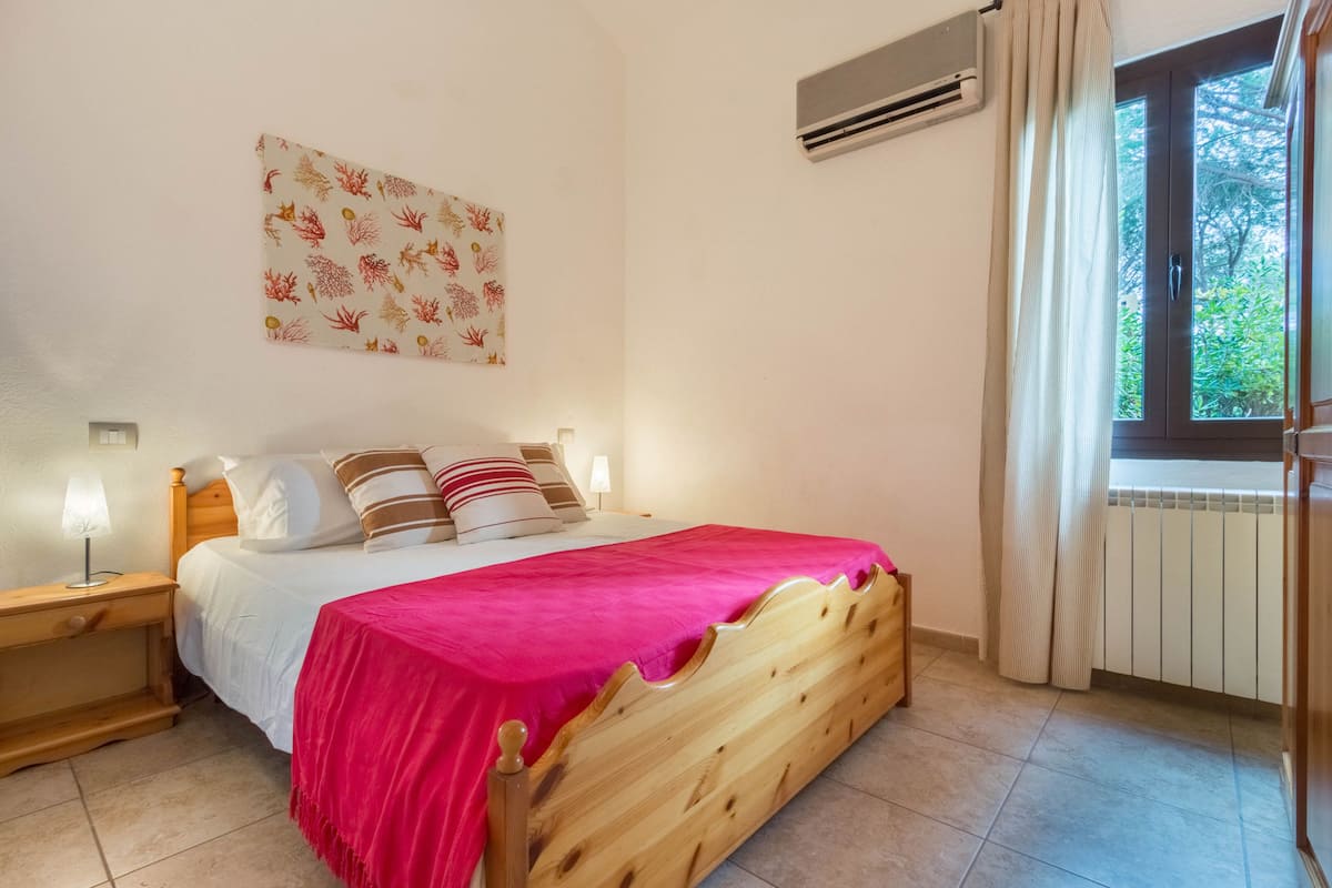 A bedroom with a wooden bed, a nightstand, a bedside lamp, a window with curtains, and a wall-mounted air conditioner.