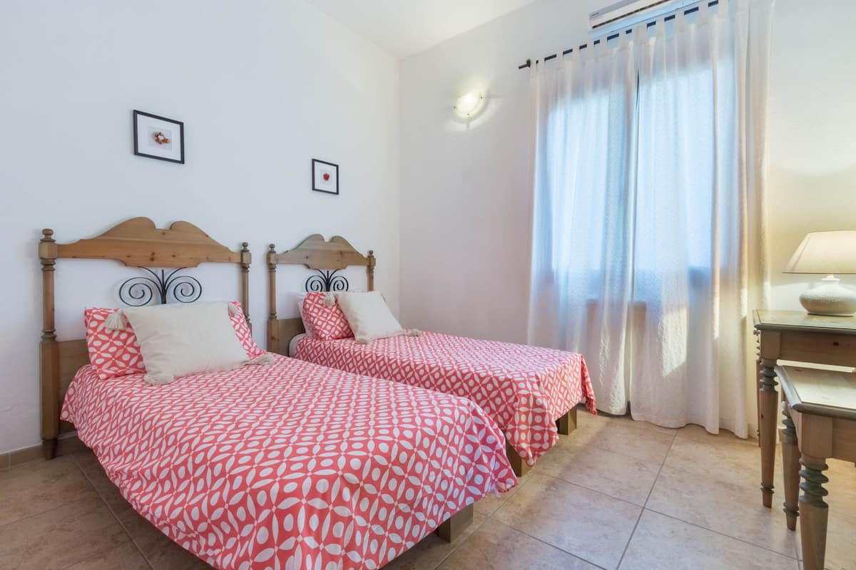 A room with two beds, each with a red and white patterned bedspread, wooden headboards, and white pillows. There is a small table with a lamp and a window with white curtains.