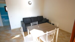 A small, bright room with a tiled floor, a wooden table, white chairs, and a black sofa. - Myrsine 2 (Olbia)
