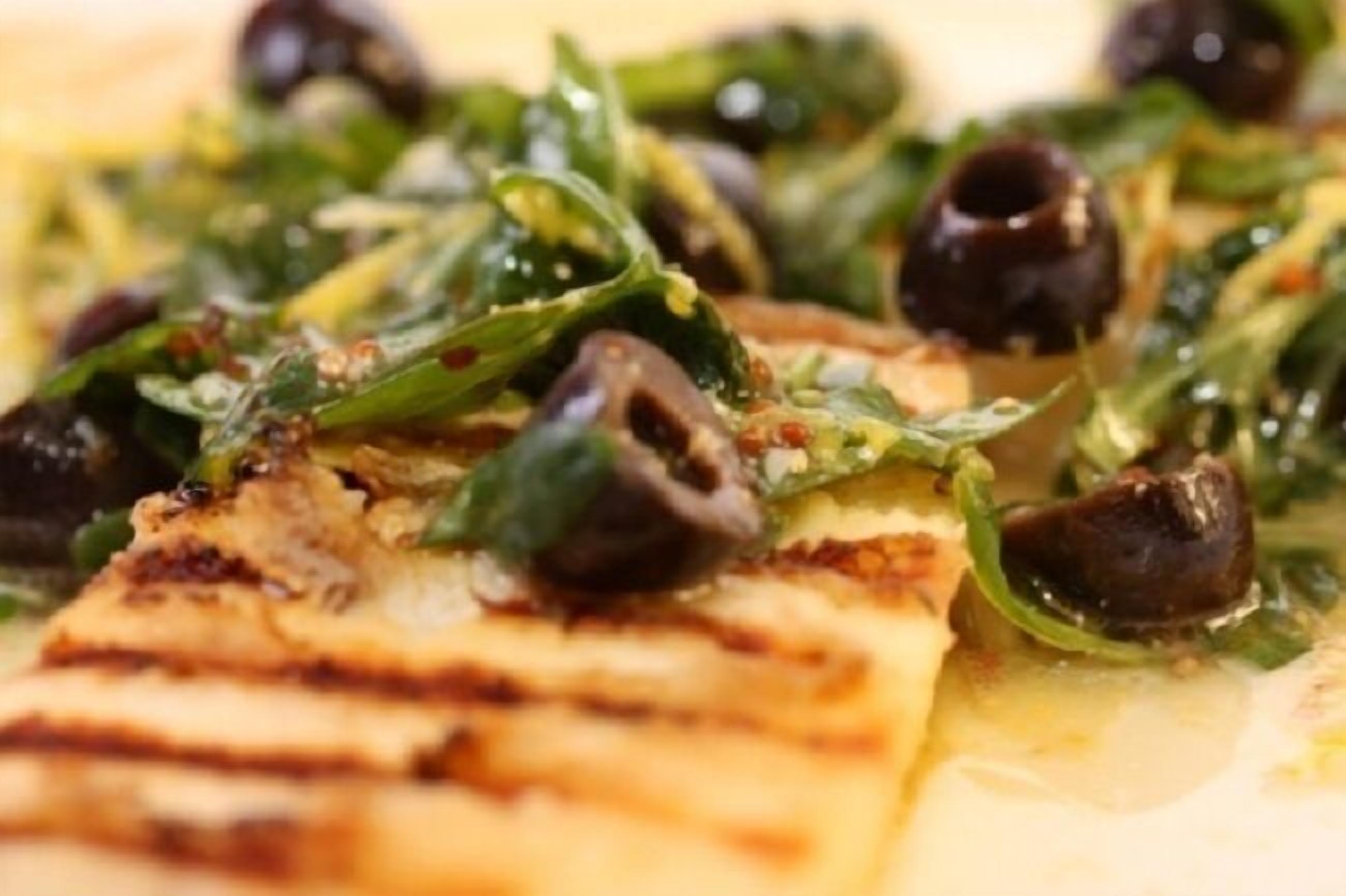 A close-up of a dish with grilled meat, greens, and black olives.
