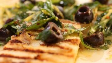 A close-up of a dish with grilled meat, greens, and black olives.