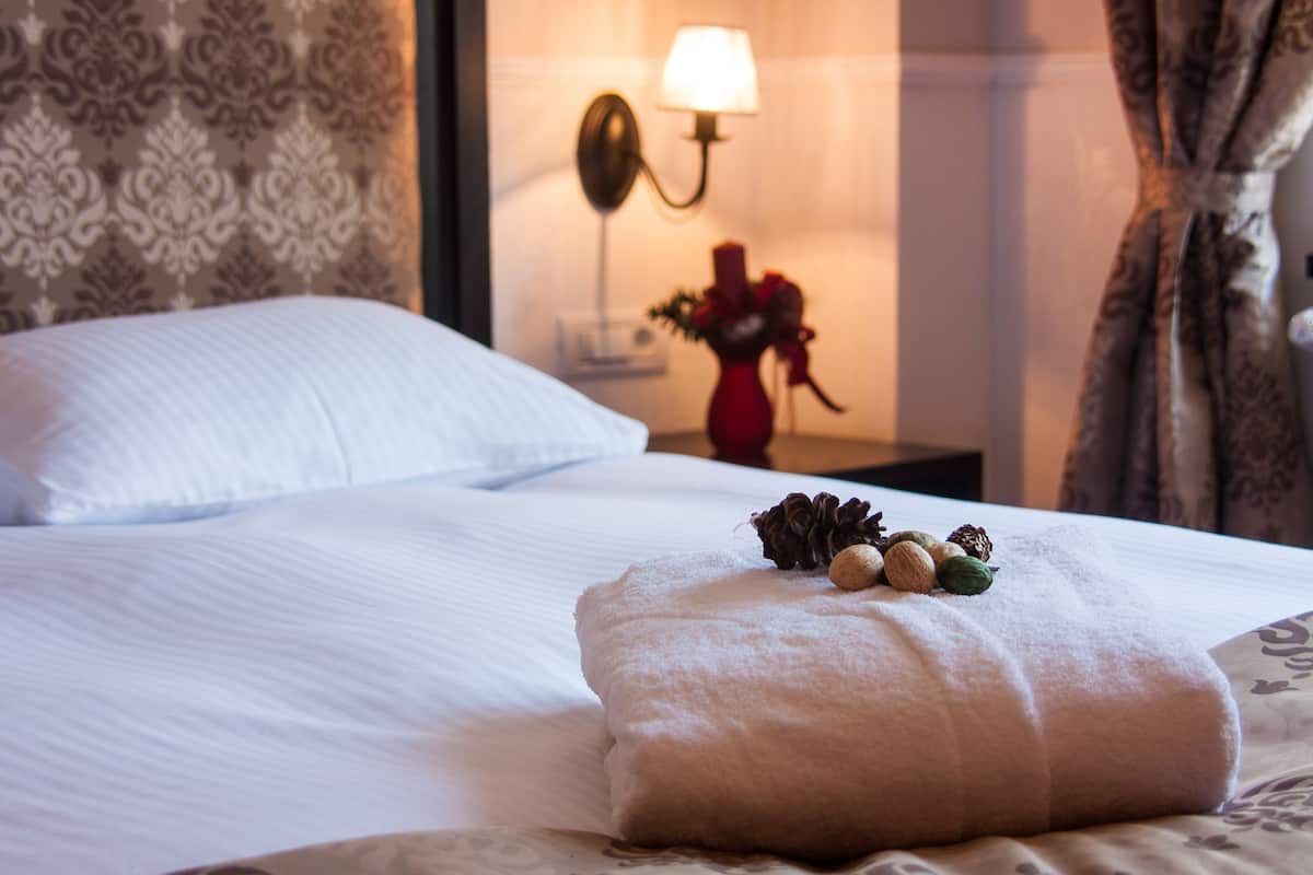 Ein ordentlich bezogenes Bett mit einem weißen Kissen und einer beigen Decke. Ein kleiner Tisch mit Lampe und Blumenvase steht daneben.