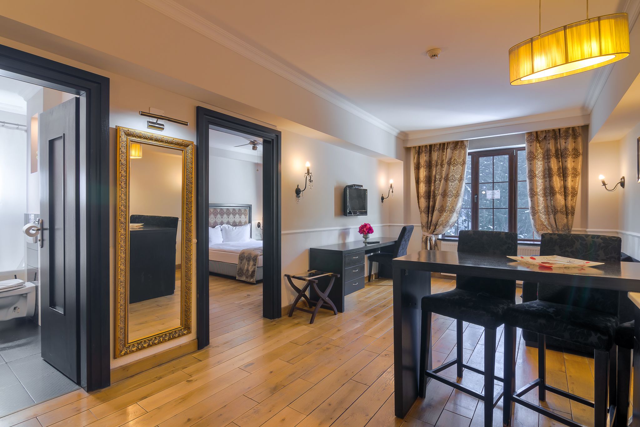 Een hotelkamer met een houten vloer, een grote spiegel, een bed, een bureau, een stoel, een televisie en een raam met gordijnen.