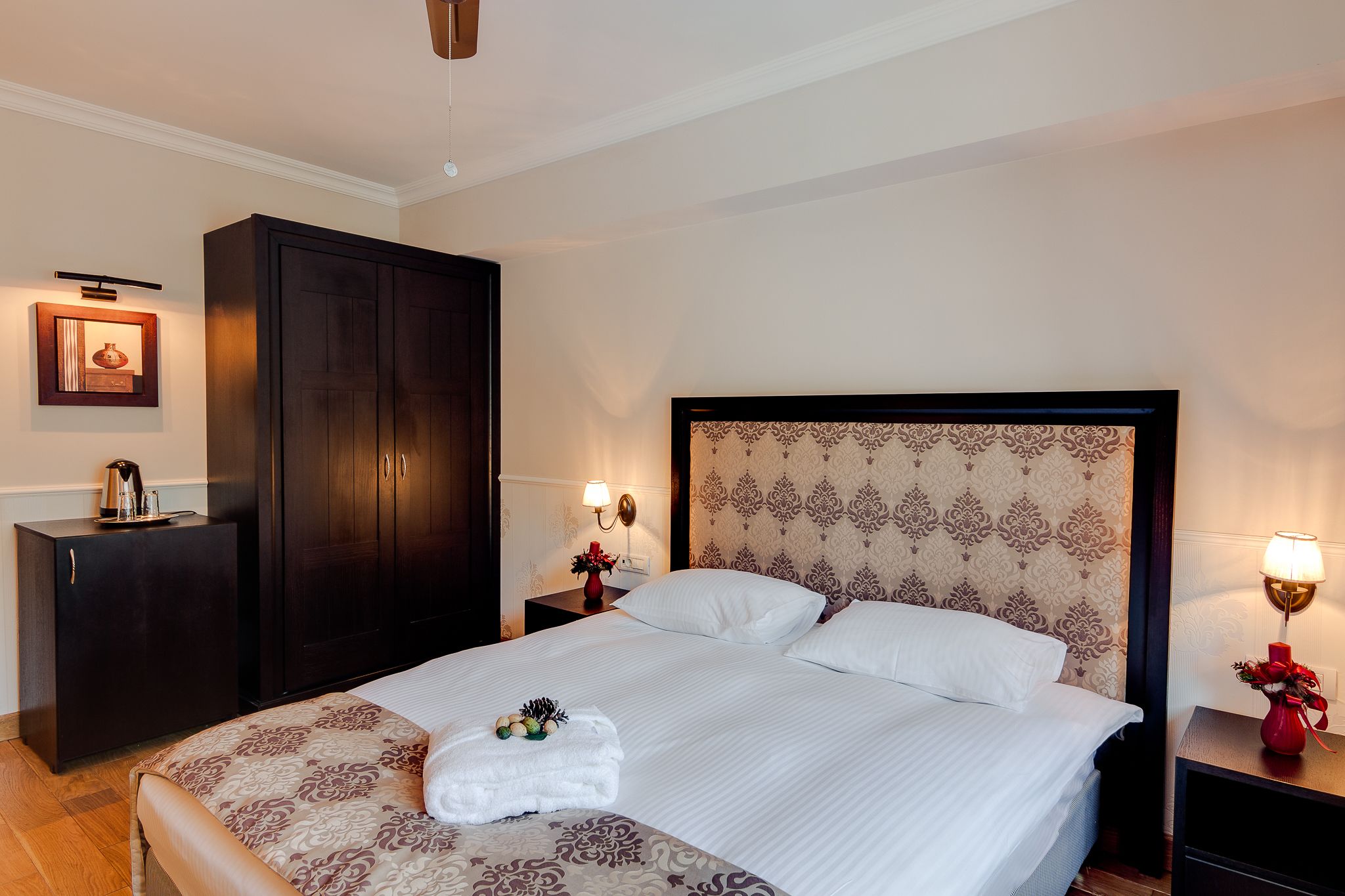 A neatly made bed with a decorative headboard, a dark wooden wardrobe, and a small table with a lamp and flowers.