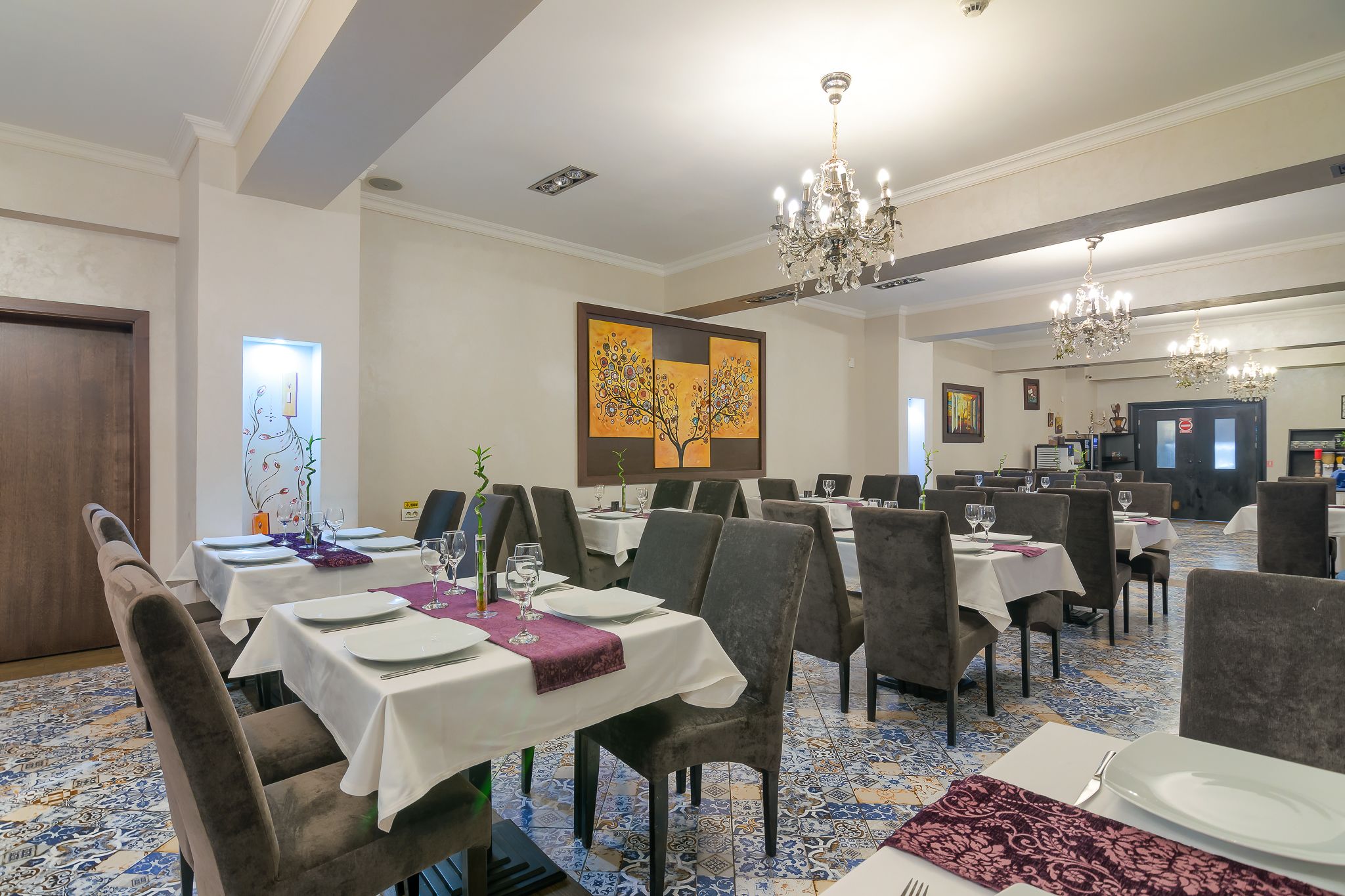 A dining area with tables set for service, a chandelier, and a large painting on the wall.
