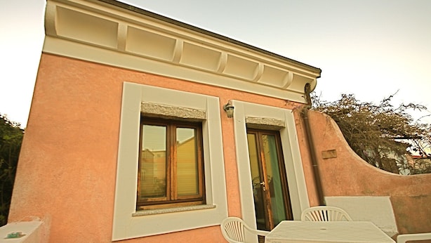 A building with a pink facade, two windows with white frames, a green door, and a small outdoor seating area with white chairs and a table. - Casa Assumptha 1p (Olbia)
