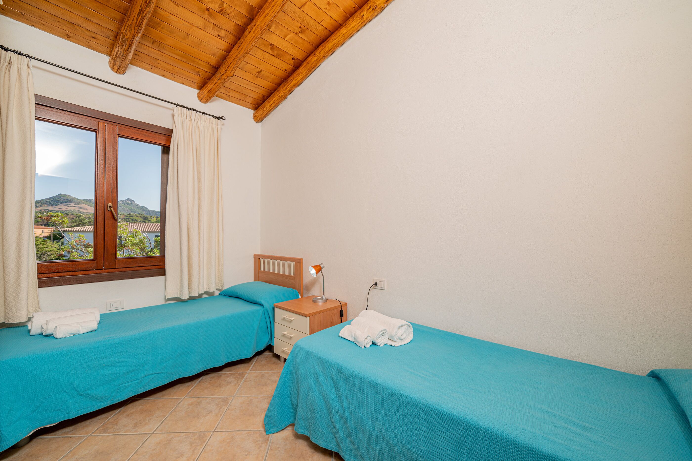 A room with two beds, a wooden headboard, a nightstand, a lamp, and a window with curtains.