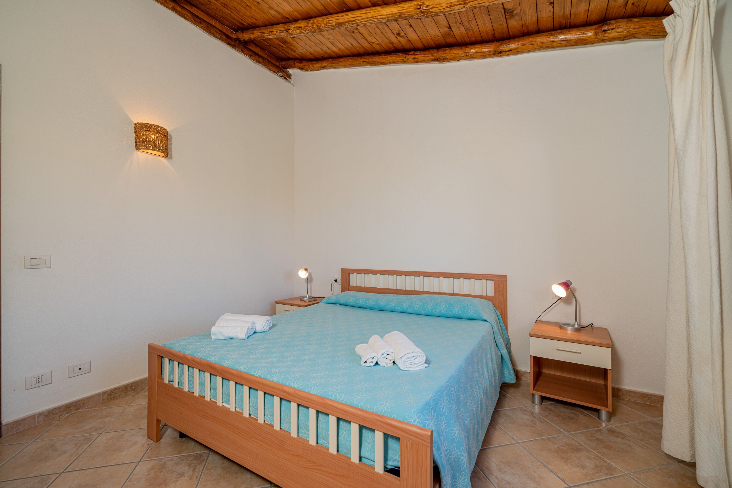 Quarto com cama de madeira, mesa de cabeceira com abajur e luminária na parede.