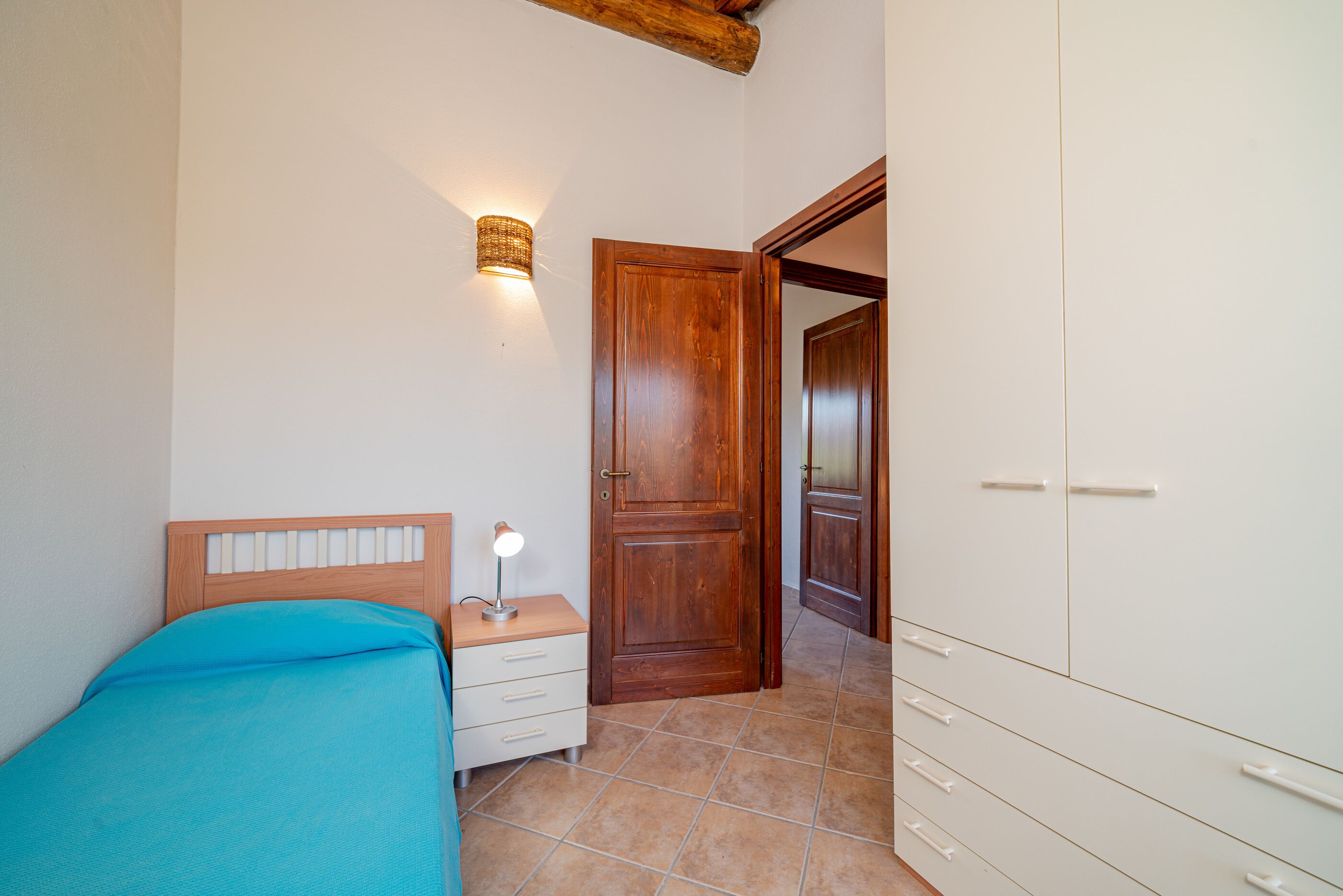 A bedroom with a bed, bedside table, lamp, wardrobe, and a door leading to another room.
