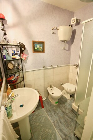 A bathroom with a sink, bidet, toilet, and a small shelf with toiletries. - Casa Serenella (Budoni)