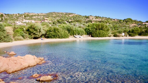 A clear coastal beach with rocky formations in the foreground and a hillside with buildings in the background. - Villa Beatrice (Olbia)