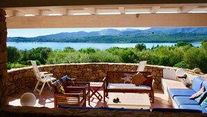 A stone-walled outdoor seating area with wooden furniture and a view of a lake and mountains. - Villa Beatrice (Olbia)