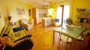A living room with a sofa, a dining table with chairs, a kitchen area with cabinets, and a television. - Casa Antonella (Olbia)
