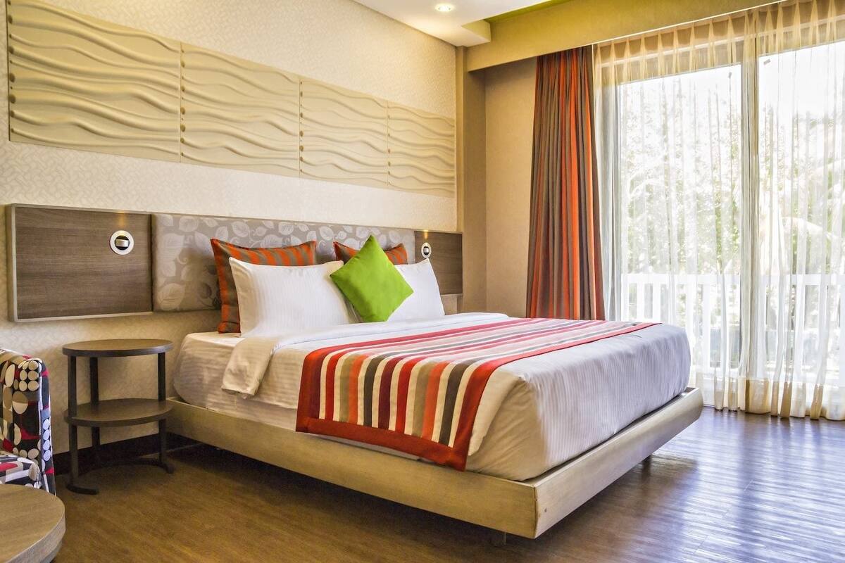 A spacious bedroom with a large bed, a bedside table, and a view of greenery through the window.
