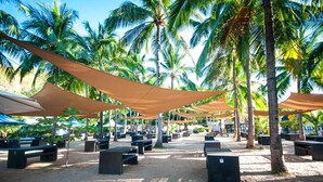 An outdoor seating area with tables and chairs under shade cloths, surrounded by palm trees. - Camaya Coast Hotel (Mariveles)