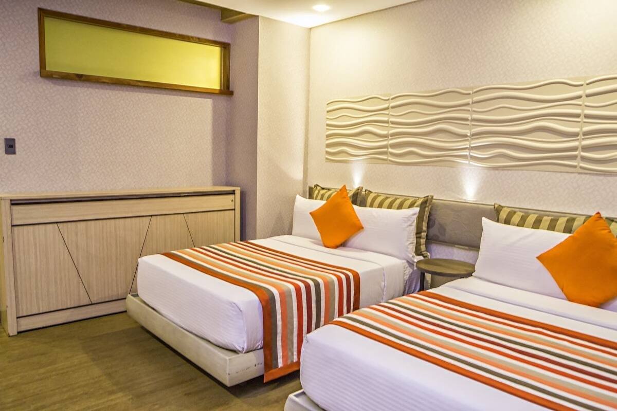 A hotel room with two beds, each with striped bedding and orange pillows. There is a headboard with a wavy design, a small bedside table, and a wall-mounted light fixture.