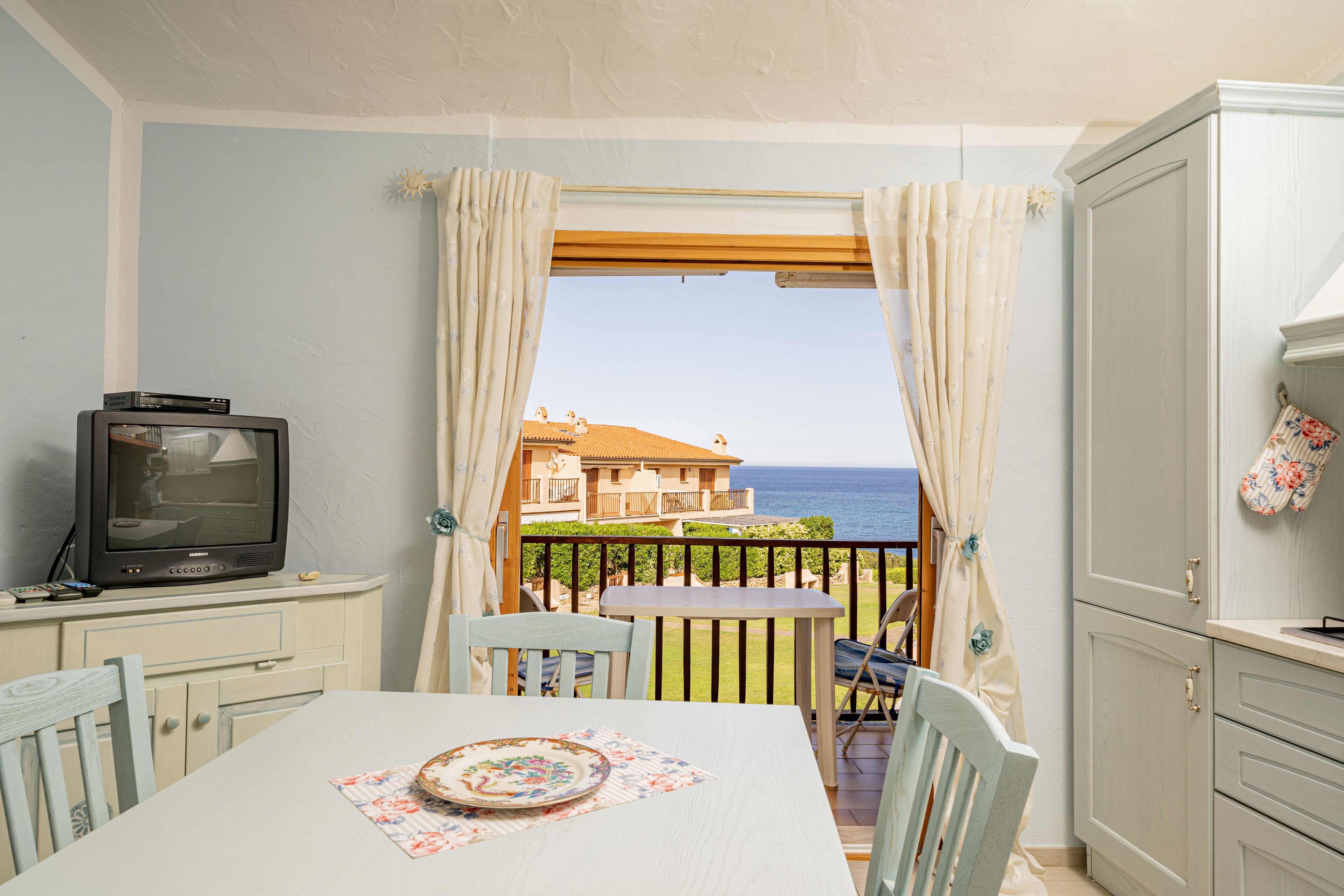 A room with a dining table, chairs, a television, and a view of the sea through a window with curtains.