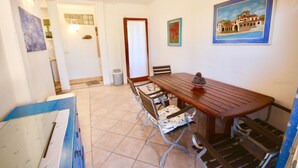 Un comedor con una mesa de madera, seis sillas y un banco. Hay dos cuadros en la pared, un cesto de basura y una puerta que conduce a otra habitación.