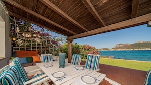 Un patio al aire libre con techo de madera, una mesa para dos, sillas a rayas y vistas al mar y a las montañas.