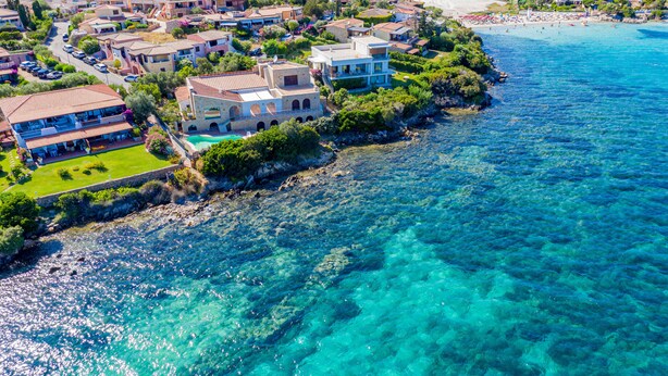 Zona residencial costera con casas cerca de un mar azul cristalino, con un edificio destacado que tiene piscina y una casa grande con balcón con vista al mar.
