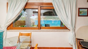 Una habitación con ventana de madera, cortinas blancas, una mesa de vidrio y una silla de madera.