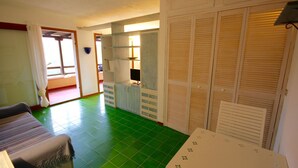 Une pièce avec un sol carrelé de carreaux verts, un lit, une table recouverte d’une nappe à motifs floraux et un ensemble d’armoires en bois à persiennes.