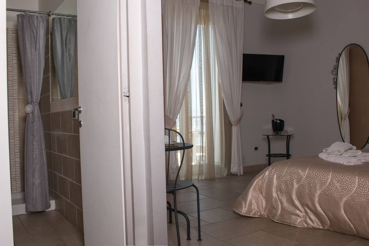 Un dormitorio con cama, televisor montado en la pared, una mesita con un cubo y baño con cortina de ducha.