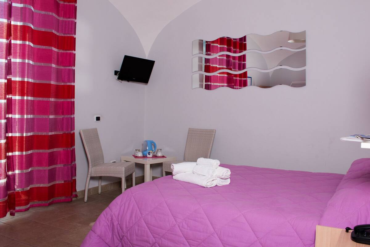 Habitación de hotel con cama, dos sillas, una mesita pequeña con un hervidor, un televisor en la pared y una mesita de noche con lámpara y teléfono.