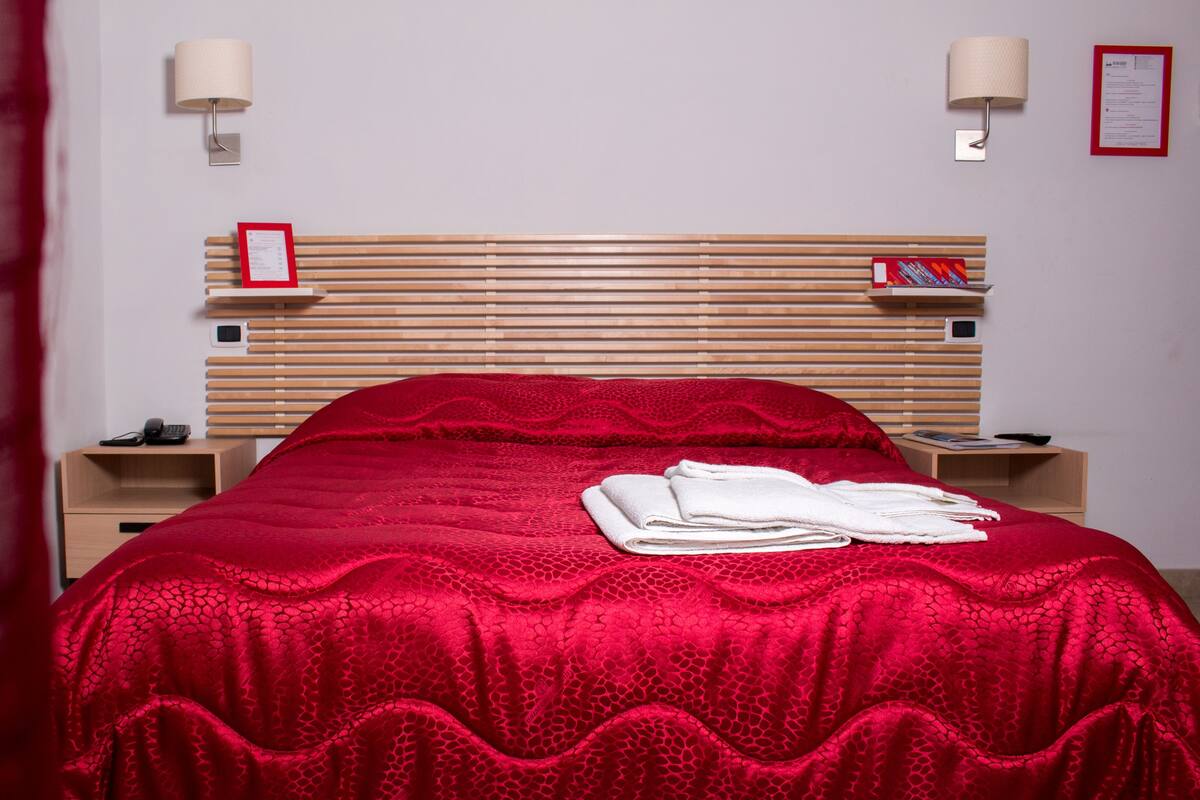 Una cama bien hecha con colcha roja y toallas blancas, cabecera de madera, lámparas de noche y una pequeña estantería con libros.