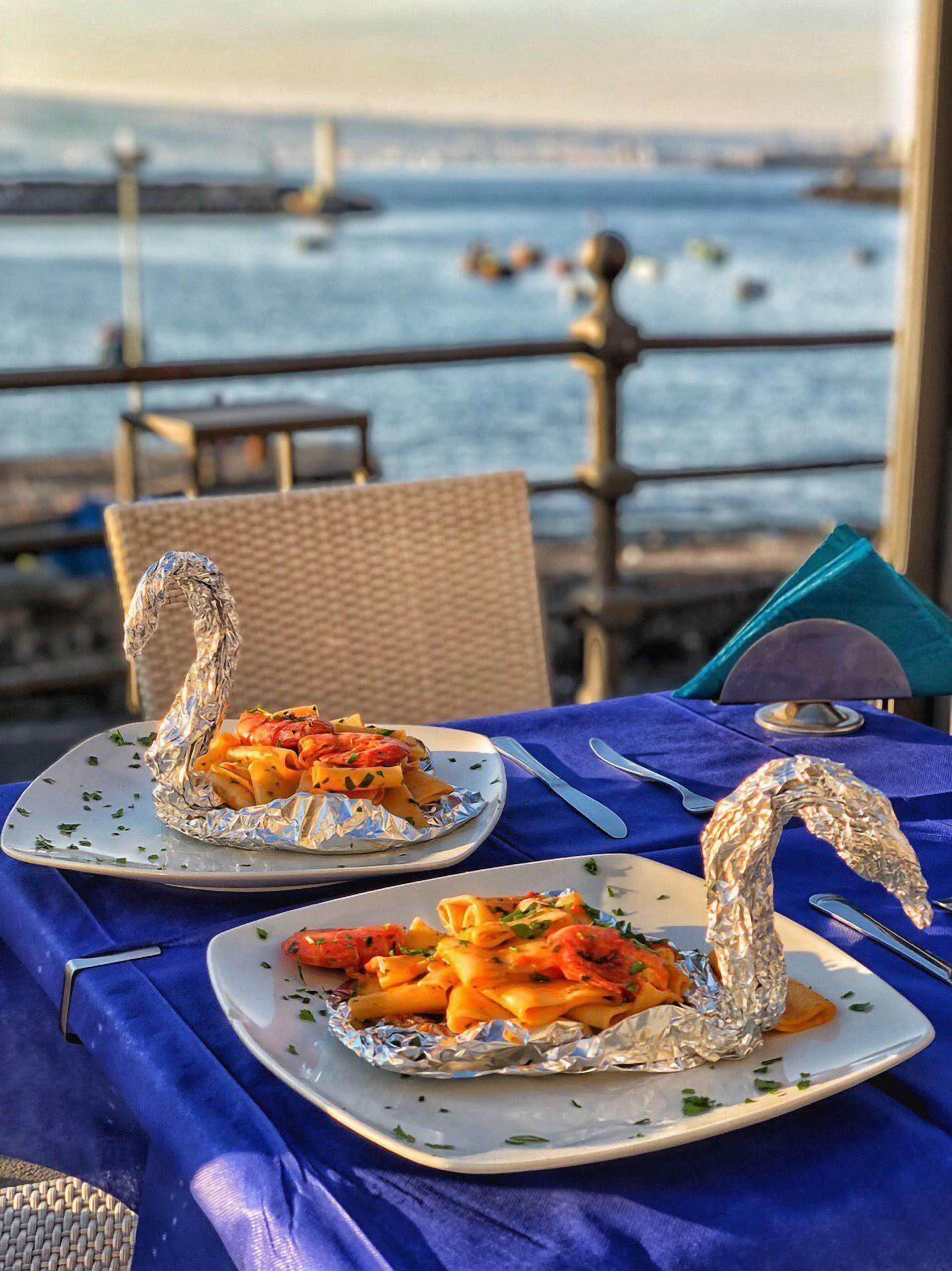 Two plates of pasta with foil-wrapped shapes on a table with a blue tablecloth, overlooking a body of water with boats.