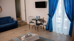 A hotel room with a sofa, a small table with chairs, a TV mounted on the wall, and a window with blue curtains. - La Dimora dei Borboni (Portici)