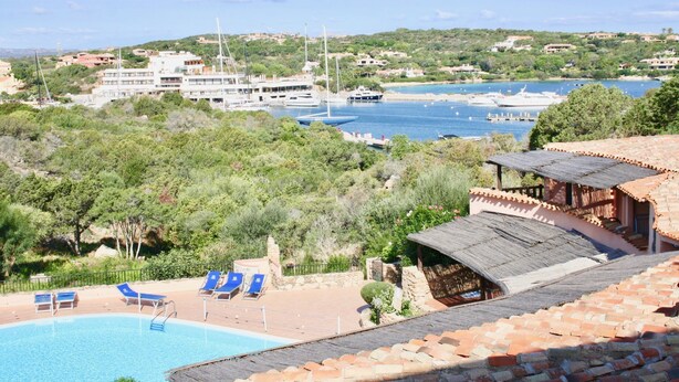 A coastal hotel with a swimming pool, lounge chairs, and a view of boats and hills.