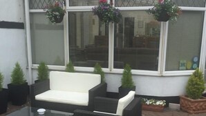 An outdoor seating area with a white sofa, two black wicker chairs, and a glass coffee table. There are hanging flower baskets above the windows. - The Old Coach House (Blackpool)