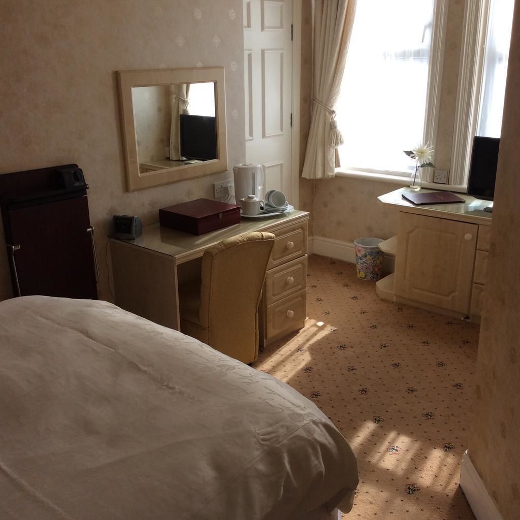 A hotel room with a bed, a desk with a mirror, a small refrigerator, a window with curtains, and a trash can.
