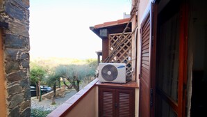 A Samsung air conditioning unit mounted on a balcony with a stone wall, wooden shutters, and a view of trees and parked cars. - Casa Rosa (Olbia)