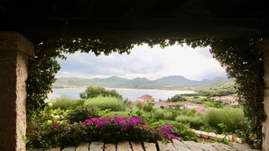 A view through a stone archway with a garden in the foreground, a body of water, and a distant village. - Casa Turchese (Olbia)