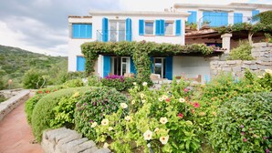 A two-story building with blue shutters, a stone wall, and a garden with various flowers. - Casa Turchese (Olbia)